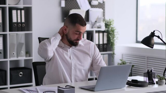 Tired businessman working online with laptop in office, massaging and stretching neck, relief muscle tension. Overload work, burnout and overload concept.
