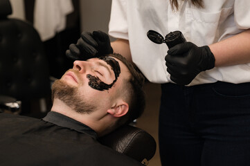 Barber applies a black moisturizing mask to a man's face while shaving. A dermatologist applies a black mask to the face of a bearded patient to clear pores and wrinkles.