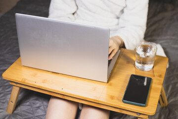 Woman is Working with laptop, smartphone and glass of water on the bed. Modern gray thin laptop on...