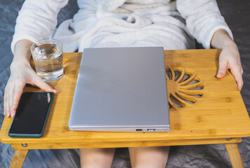 Woman is Working with laptop, smartphone and glass of water on the bed. Modern gray thin laptop on the wooden Cooling pad for laptop. Notebook Cooler for Computer, USB Fan.