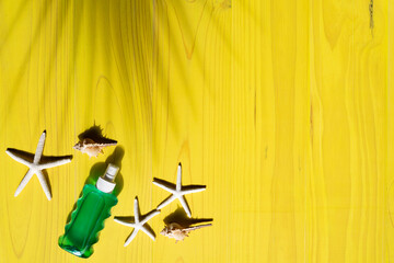 Simple composition created with a yellow background with wooden slats with visible grain and shadow of palm tree with sunscreen, starfish and conch shells and copy space
