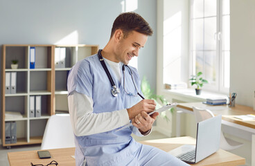 Portrait of a young male doctor or nurse in blue medical uniform sitting on the desk and working in...