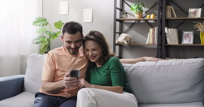 Smiling couple watching videos on phone, using social media app, planning vacation, booking hotel sitting on couch in the living room. Happy man and woman shopping online together