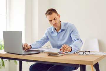 Fototapeta na wymiar Portrait of a young focused serious attractive business man working on a laptop at the desk on workplace in office and making calculations, considering new projects, analyzing company.