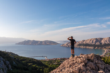 Hiker man on edge of rock cliff overlooking archipelago of Kvarner Bay in coastal town Baska, Krk...