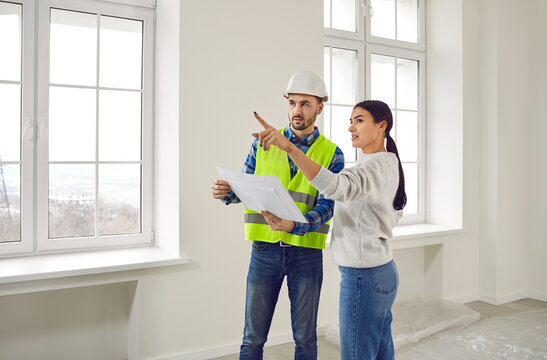 Meeting female homeowner and builder in living room for further renovation work.Attentive foreman in helmet and overalls with document or technical plan in his hands communicates with hostess of house