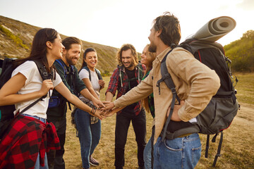 Group of friends having fun on hiking trekking holiday in countryside. Team of happy male and female hikers with rucksack bags stacking hands standing in circle on rural field with hills in background