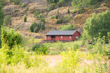 red wooden house at the foot of Norwegian mountain fjords among green trees on a sunny summer day.