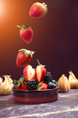 Falling strawberry into porceline pot with mix berries on dark marble background.