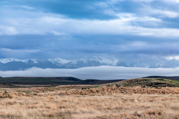 Photograph of a snow-capped mountain range running behind a large brown agricultural field with low level grey clouds on the South Island of New Zealand