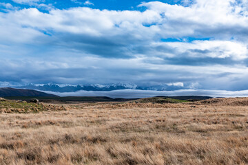 Photograph of a snow-capped mountain range running behind a large brown agricultural field with low level grey clouds on the South Island of New Zealand