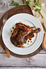 white plate with a roasted chicken with honey and soy sauce decorated. It is on a wooden table with a tea towel and a slice of bread. You can also see a parsley leaf.