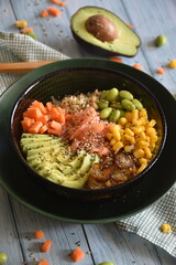 a black bowl of arooz poke, a typical asian dish. it is served with salmon, avocado, corn, carrot, edamame and onion. It is on a wooden table with food all around. 
