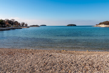 Panoramic view of small islands seen from pebble beach in coastal town Funtana, Istria, Croatia. Calm sea surface of Adriatic Mediterranean Sea in morning hours. Seaside vacation concept in summer