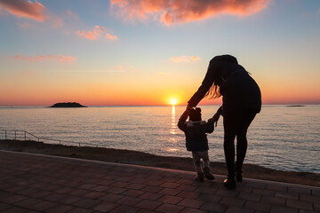 Silhouette of loving mother with small toddler walking on idyllic beach at sunset in coastal town...
