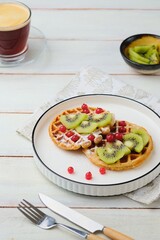 Two round waffles made of buckwheat flour, dusted with powdered sugar, topped with chopped fresh kiwi and red currants on a white plate on a light wooden background. Waffle recipes. Breakfasts.