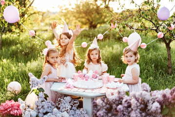 Summer outdoor kids birthday party. Group of happy Children celebrating birthday in park. Children blow candles on birthday cake. Kids party lilac pink pastel decoration and food. Presents and sweets.