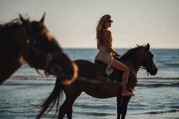 An adventurous tattooed lady is horseback riding at the beach on sunset.