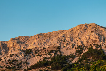 Fototapeta na wymiar Panoramic view during sunrise of majestic mountains with massive rock formation in coastal town Baska, Krk Otok, Primorje-Gorski Kotar, Croatia, Europe. Illuminated mountain peak in soft red colours.