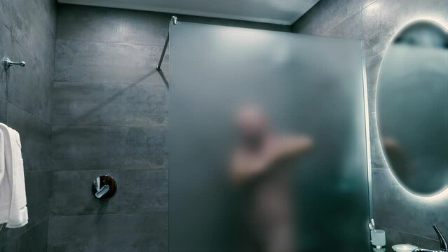 An adult man is washing his underarms behind a frosted glass wall in the shower room.