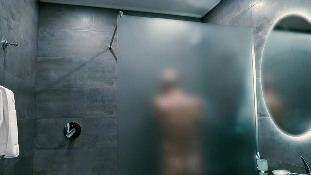 A middle-aged Caucasian man is taking a shower behind a privacy glass wall in a contemporary bathroom setting.