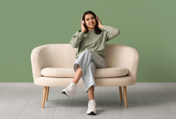 Beautiful young happy woman in headphones sitting on comfortable sofa and listening music near green wall