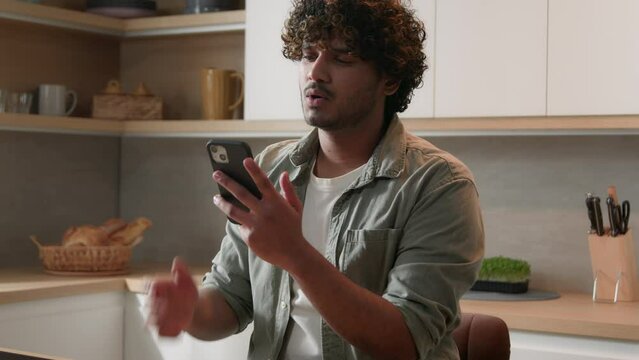 Indian Arabian man at home kitchen playing online game betting cheering favorite sport team with mobile phone unhappy failure upset bad result loose. Male guy with smartphone lost internet connection