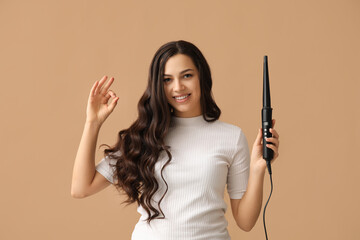 Beautiful young woman with curling iron showing OK on beige background