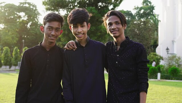 A young group of three happy Indian Asian teenager friends wearing ethnic black kurtas or Pathani standing together outdoors with hands on each other's shoulders looking or facing the camera smiling 