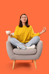 Fototapeta na wymiar Young woman with cup of tea meditating in armchair on orange background