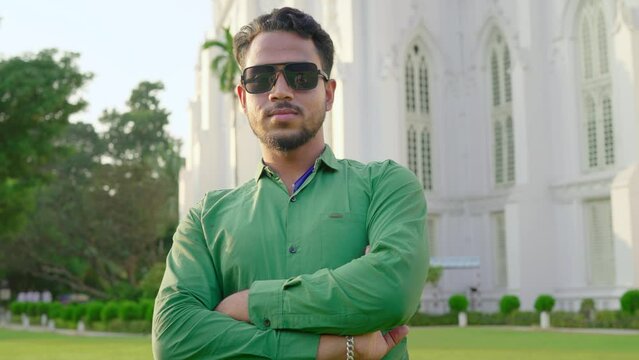 A young cool hipster confident Indian Asian man or male with crossed arms and wearing sunglasses standing outdoors in front of the church or white building looking or facing the camera on a sunny day 