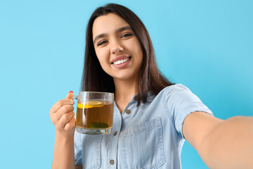 Young woman with glass cup of lemon tea taking selfie on blue background, closeup