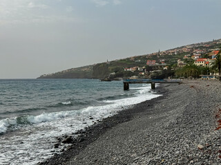 Scenic view of idyllic black pebble beach Palmeiras at Santa Cruz, Madeira island, Portugal, Europe. Looking at majestic Atlantic Ocean on overcast cloudy day. Serene tranquil atmosphere at promenade