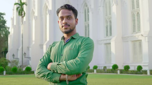 A young confident Indian Asian male or man is standing outdoors with crossed arms in front of a white building looking or facing the camera smiling on a sunny day 