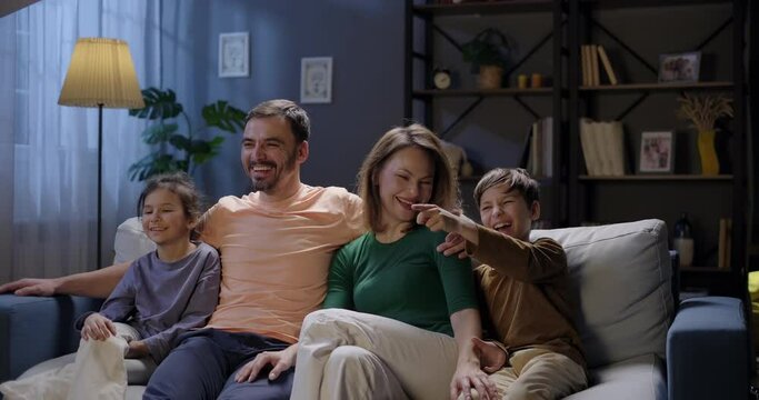 Full family watching TV funny movie or show, laughing sitting on the couch in the living room in the evening. Couple with children watching television streaming service together. Family leisure time