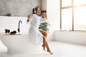 Cheerful black woman in bathrobe singing joyfully with smartphone in bathroom