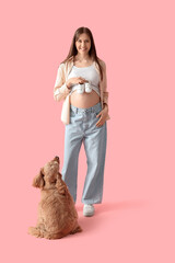 Young pregnant woman with baby booties and cute cocker spaniel on pink background