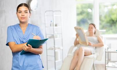 Fototapeta premium Woman doctor holds folder with papers and enters medical information about client of clinic into form. In background, female visitor is lying in armchair and looking at reflection in small mirror