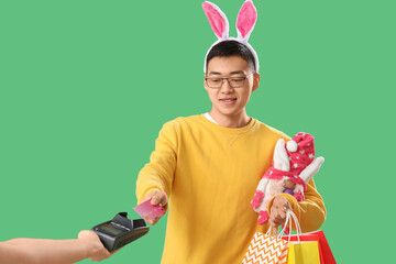 Young Asian man in bunny ears with shopping bags paying via terminal on green background. Easter...