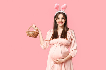 Beautiful young pregnant woman in bunny ears with wicker basket on pink background. Easter...