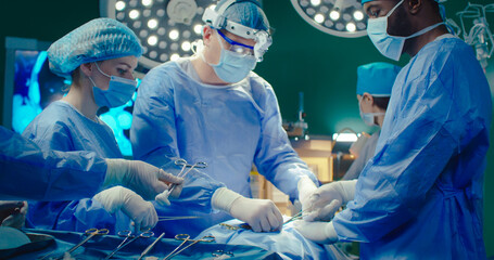 Diverse team of professional surgeons performs invasive surgery on patient in hospital. Nurse gives...