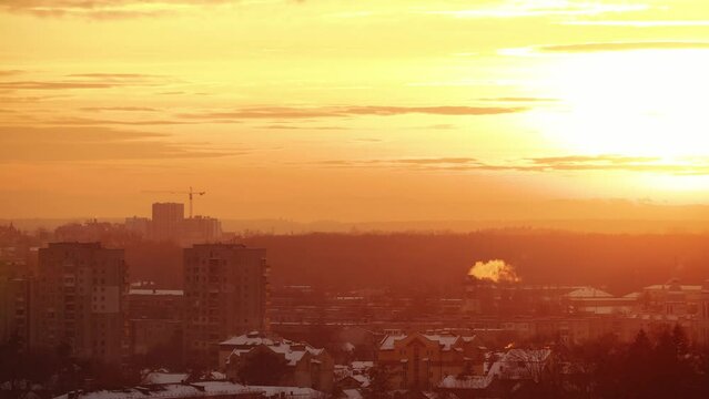 A golden sunset bathes a wintry cityscape, with smoke rising from a distant stack against a tranquil evening sky.