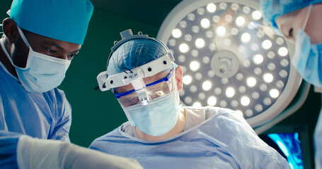 Close-up of three doctors dressed up in special clothes. Male doctor in binoculars. African American in special mask and gloves asssists operation. Medical workers at background of operating lamp.