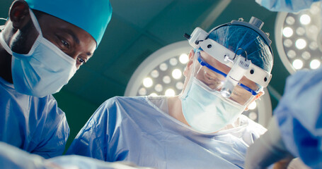 Shoot from below. Close-up medical workers during surgery intervention. African american intern...