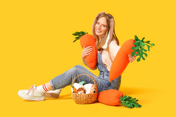 Beautiful woman in bunny ears with toy carrots and Easter basket sitting against yellow background