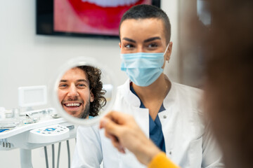 Young adult happy male patient feeling confident with a new smile sitting with a female dentist after successful treatment in a modern dental clinic.