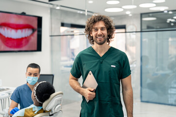 Handsome dentist in green uniform holding digital tablet in modern clinic with patient in dentist's chair and nurse in background. Healthy smile presentation on tv screen also visible in background.