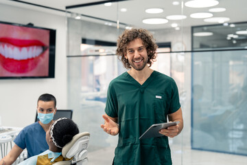Highly qualified young dentist holding fintech device at modern clinic with patient in dentist's chair and nurse in background. Hollywood smile presentation on tv screen also visible in background.