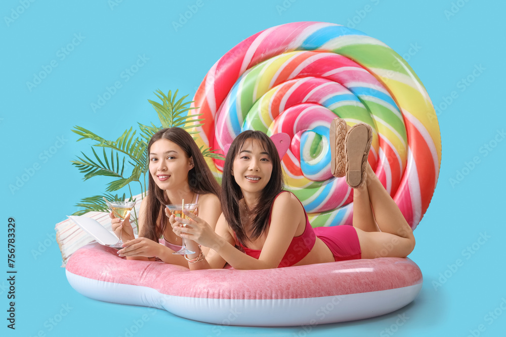 Sticker Female Asian friends with cocktails lying on swim mattress against blue background