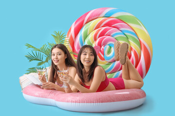 Female Asian friends with cocktails lying on swim mattress against blue background - obrazy, fototapety, plakaty